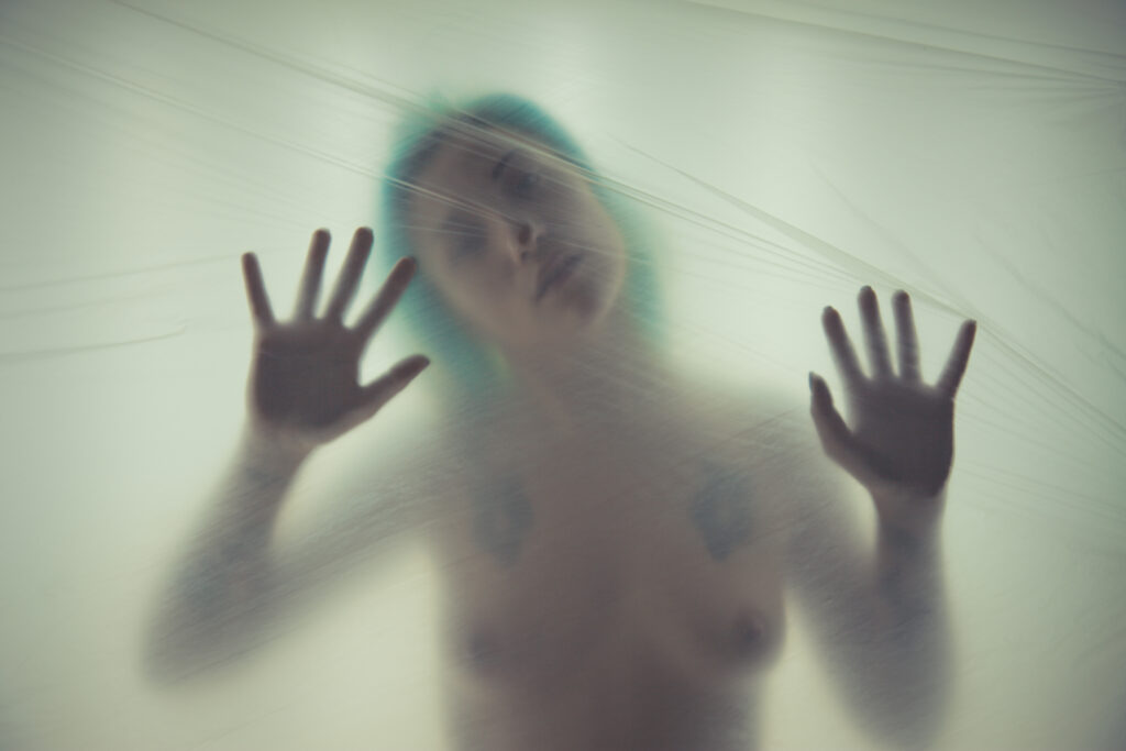 Foto artistica, la modella è dietro un telo trasparente, le mani sono appoggiate al telo, il capo è inclinato di lato, si intravedono i lineamenti dei capelli, degli occhi e delle labbra, così come il busto, il corpo è nudo; solo il naso è appoggiato al telo come le mani. La figura è sfocata, dona un senso onirico.